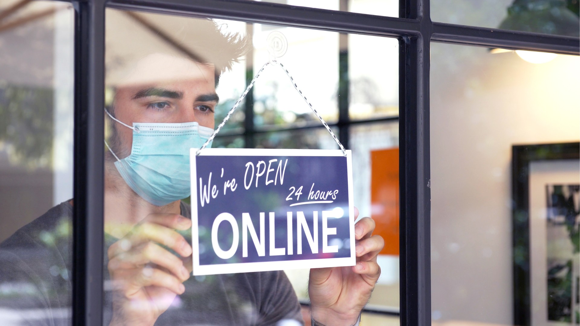 Mann mit Mundschutz hängt Schild an die Tür: 24 Stunden online geöffnet 